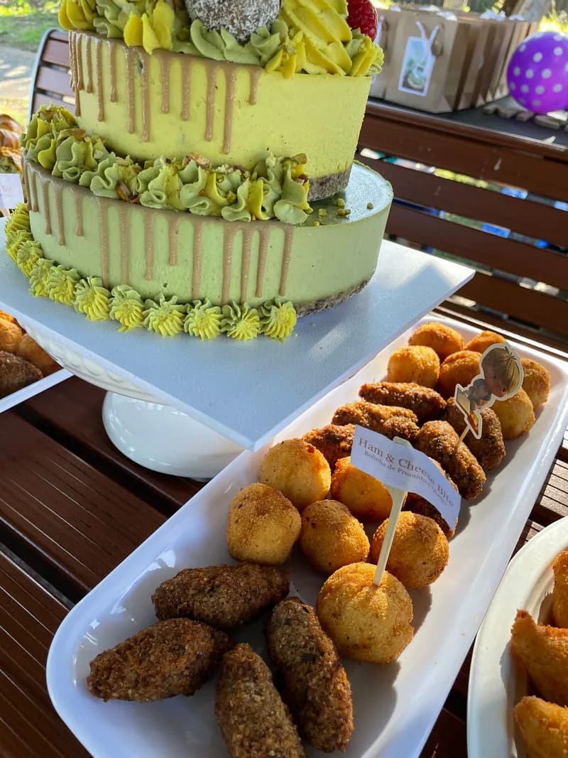 A pistachio birthday cake beside a platter of quibes and bolinhas with named picks