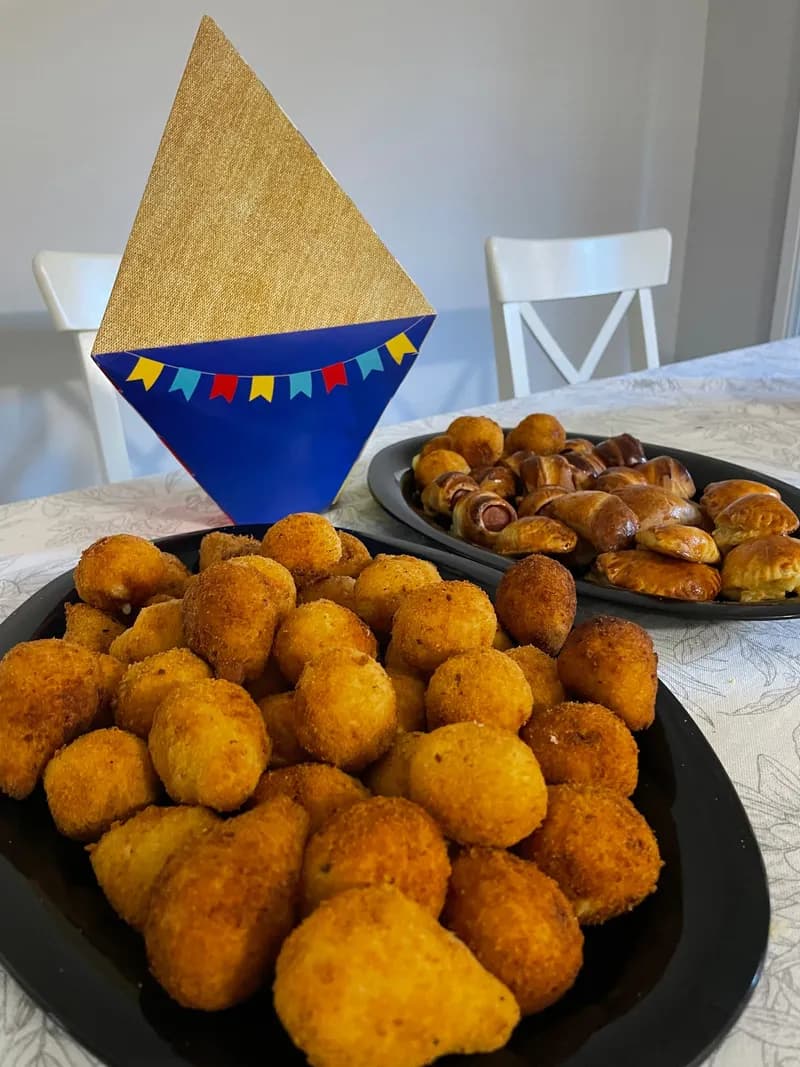 A black bowl heaped with bolinhas de queijo beside sausage rolls, with a Festa Junina hat