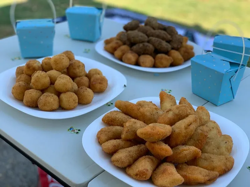 Three plates of coxinhas, quibes and risoles on a picnic table on the grass, with takeaway boxes